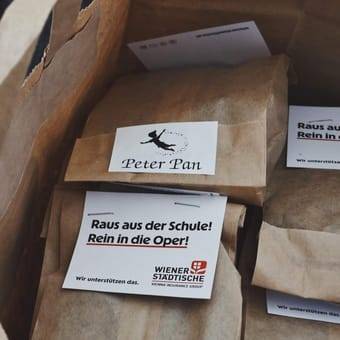 Mehrere braune Papiertüten mit verpackten Keksen; auf einer Tüte ist ein Aufkleber mit „Peter Pan“ und auf einer Karte steht „Raus aus der Schule! Rein in die Oper!“ mit dem Logo der Wiener Städtische.
