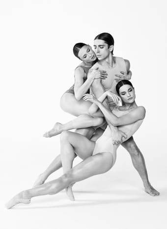 Schwarz-Weiß-Foto von drei Tänzerinnen in enger, ineinander verschlungener Pose; ruhig und ausdrucksstark vor hellem Hintergrund.